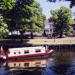 Zabytkowa łódź kanałowa pływająca w słoneczny dzień w Norwich w Wielkiej Brytanii, z malowniczymi domami i drzewami wzdłuż brzegu rzeki.