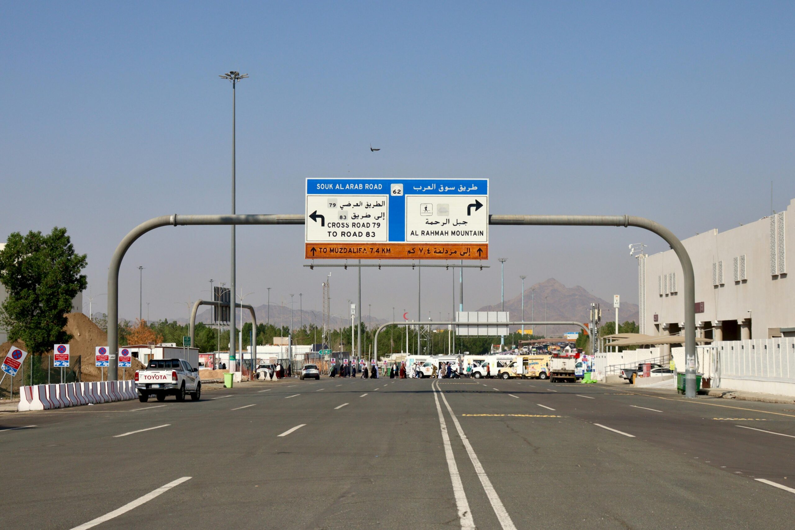 Oznakowanie drogowe prowadzące do góry Al Rahmah w Mekce, z pojazdami i ludźmi w słoneczny dzień.