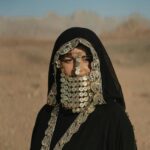 Kobieta ubrana w tradycyjny strój beduiński z biżuterią w postaci monet w pustynnym krajobrazie.