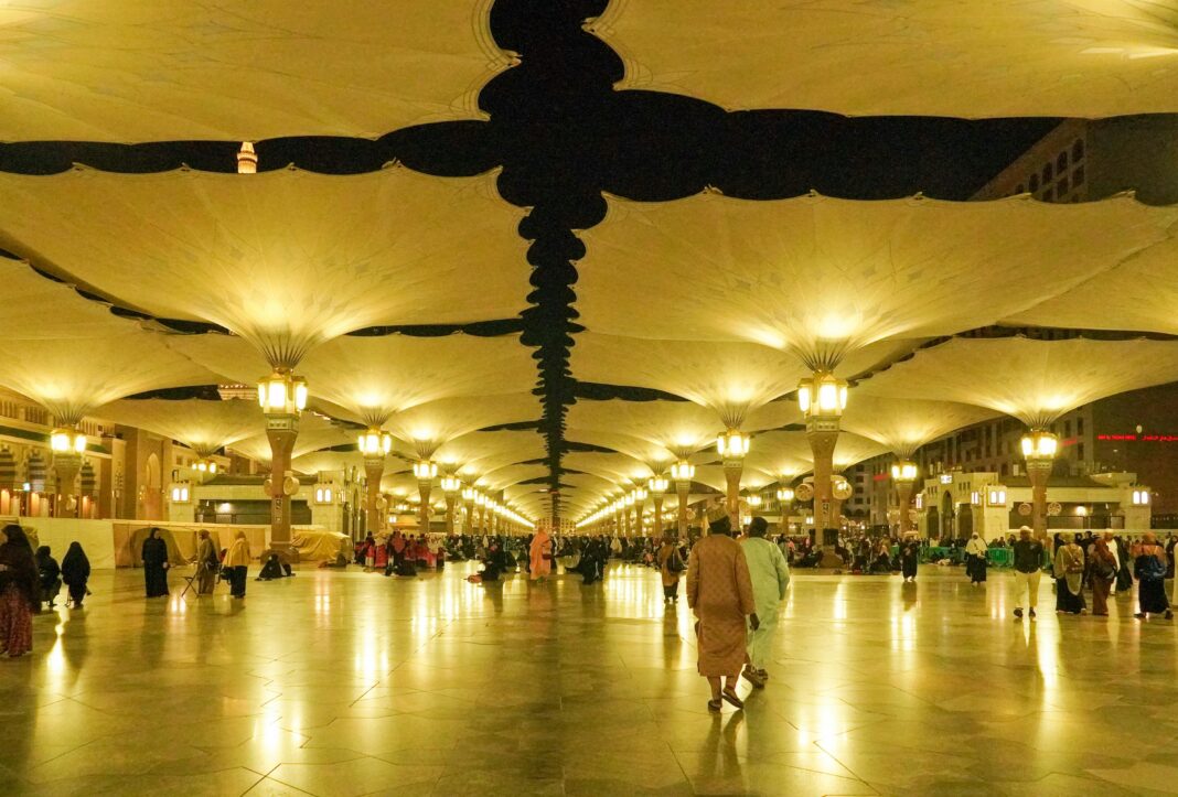 Oświetlona scena nocna na dziedzińcu Masjid Nabawi w Medynie w Arabii Saudyjskiej, przedstawiająca architekturę i parasole.