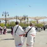 Pielgrzymi w tradycyjnych strojach spacerujący na świeżym powietrzu w Medynie w Arabii Saudyjskiej z parasolami w tle.