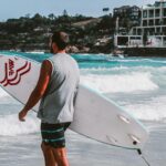 Mężczyzna trzymający deskę surfingową na plaży Bondi w Sydney, gotowy do surfowania po tętniących życiem falach.