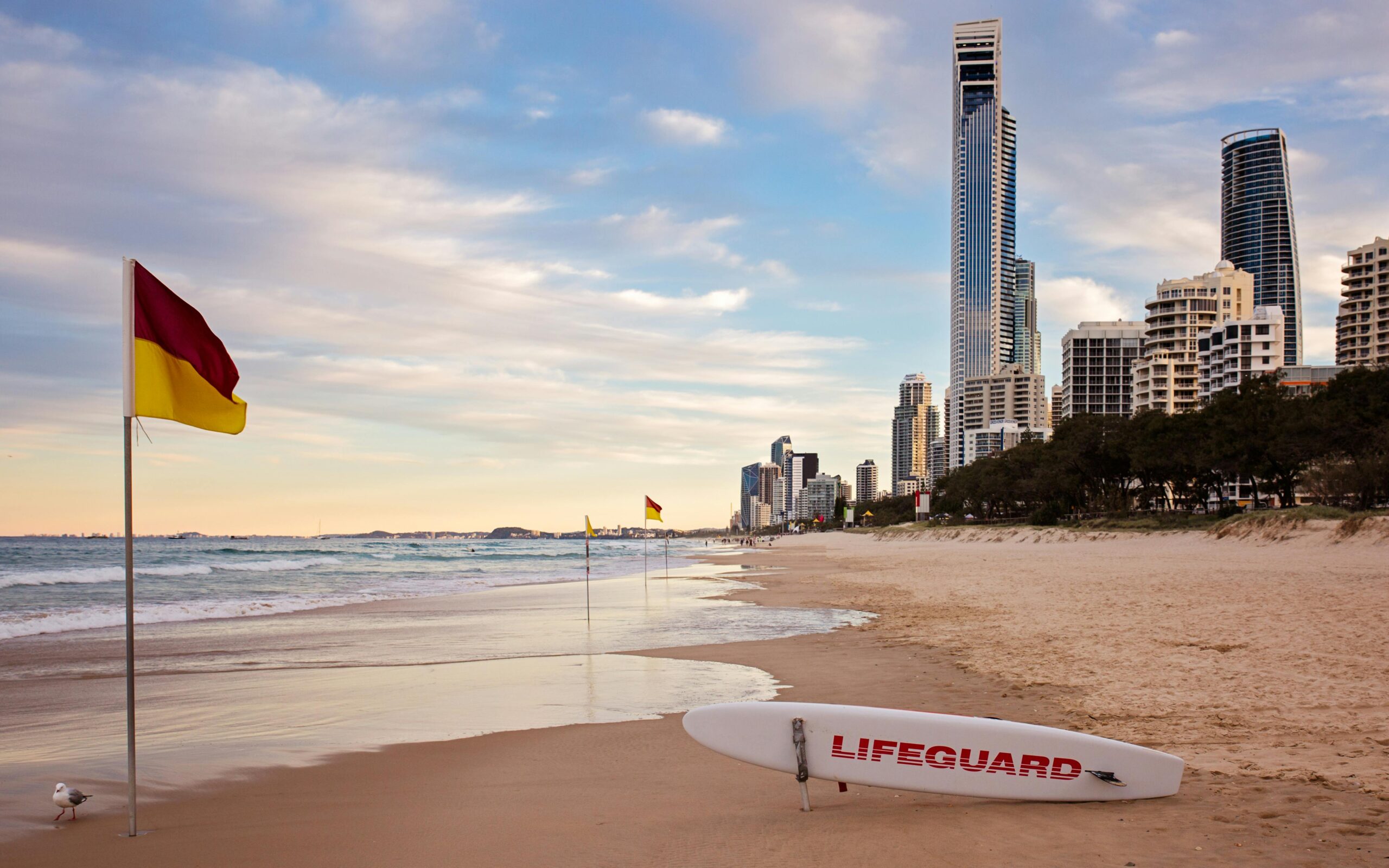 Ujęcie plaży Surfers Paradise z panoramą o zachodzie słońca, widoczną tablicą ratownika i flagami.