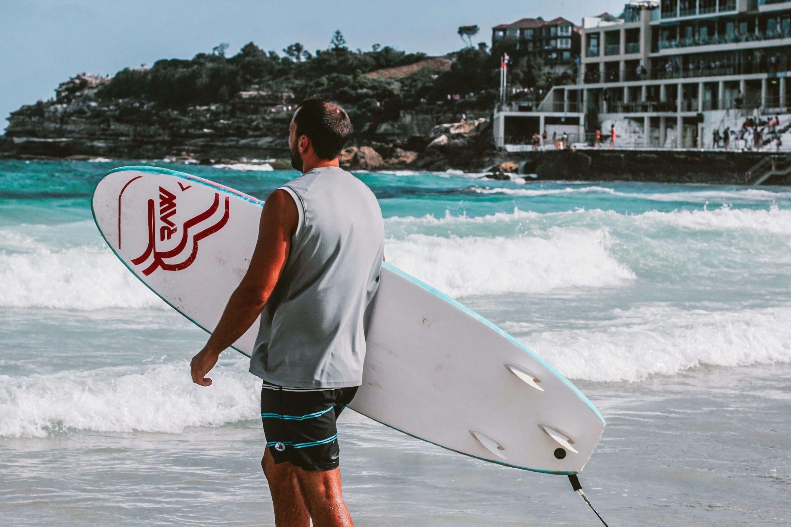Mężczyzna trzymający deskę surfingową na plaży Bondi w Sydney, gotowy do surfowania po tętniących życiem falach.