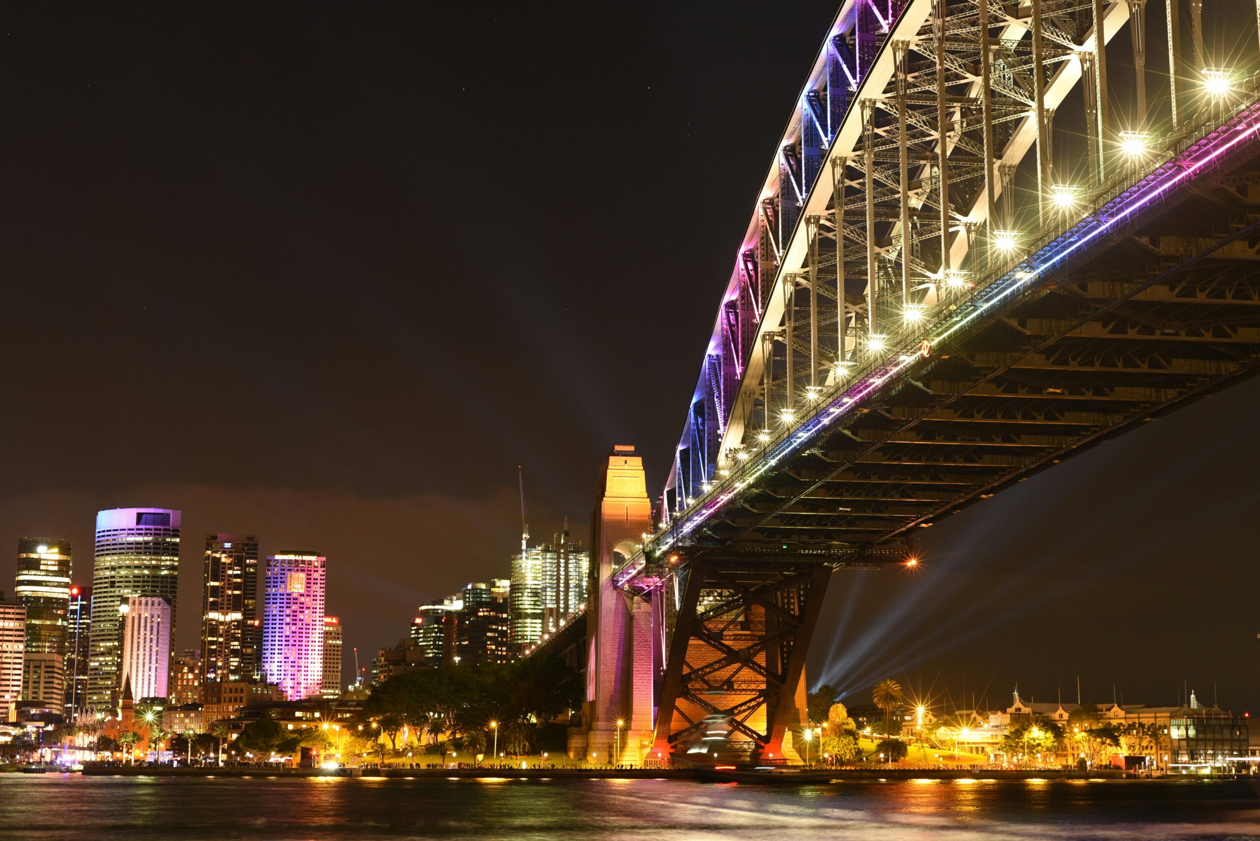 Oszałamiający nocny widok na most Sydney Harbour Bridge z oświetloną panoramą miasta odbijającą się w wodzie.