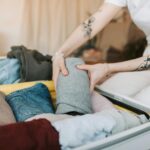 Co spakować do bagażu podręcznego na wyjazd służbowy? Zbliżenie osoby pakującej ubrania do walizki, pokazującej tatuaże na ramionach, w przytulnym pomieszczeniu.