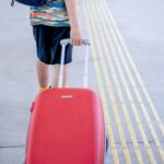 Osoba z czerwonym bagażem spacerująca po słonecznym chodniku na świeżym powietrzu, chwytająca chwilę podróży.