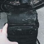 Ręka sięga po plecak w kamuflażu w pobliżu koła rowerowego, idealny do podróży i przygód.