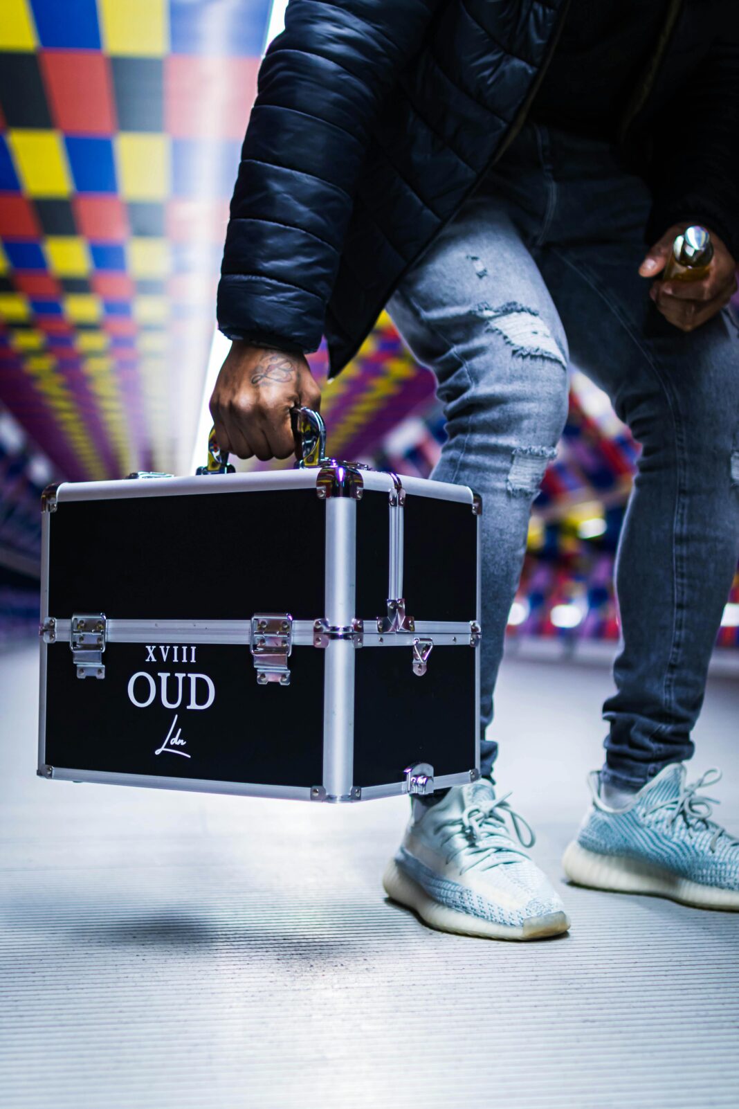 Mężczyzna w tętniącym życiem korytarzu trzyma czarną metalową walizkę z napisem „XVIII OUD”.