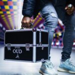 Mężczyzna w tętniącym życiem korytarzu trzyma czarną metalową walizkę z napisem „XVIII OUD”.