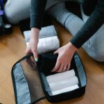 Historia bagażu podręcznego – jak zmieniały się przepisy? Osoba pakująca zwinięte ręczniki do torebki na drewnianej podłodze, przygotowująca się do podróży.