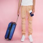 Modny młody dorosły trzymający walizkę i paszport, gotowy do podróży, na różowym tle.