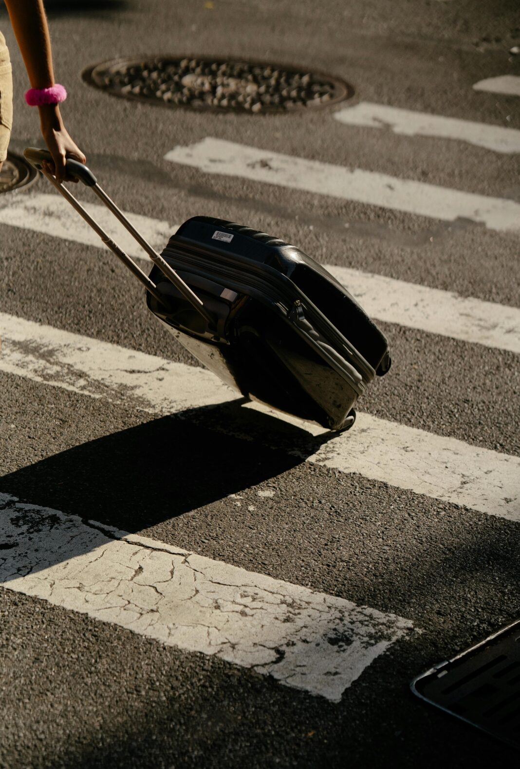 bagaz-podreczny Osoba ciągnąca walizkę przez przejście dla pieszych w słoneczny dzień.