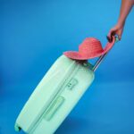 Miętowozielona walizka z różowym kapeluszem przeciwsłonecznym na niebieskim tle, przywołująca klimat letnich podróży.