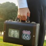 Zbliżenie dłoni trzymającej walizkę retro z naklejką Route 66 w nasłonecznionym otoczeniu na zewnątrz.