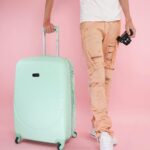 Dorosły trzyma miętową walizkę i aparat, gotowy do podróży na różowym tle.