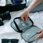 Bagaż podręczny na wyjazdy narciarskie – jak spakować się sprytnie? Zbliżenie na niezbędne w podróży przybory, w tym okulary przeciwsłoneczne i organizer na elektronikę, leżące na łóżku.