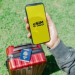 Podróżnik w Los Angeles trzymający smartfon z eSIM w pobliżu czerwonej walizki na trawie.