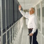 Najlepsze organizery do bagażu podręcznego Widok z boku starszego, spokojnego mężczyzny w formalnym ubraniu z walizką spacerującego nowoczesnym korytarzem lotniska i machającego na pożegnanie, patrząc przez szklaną ścianę