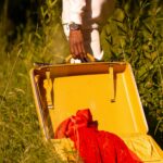 Osoba stojąca z otwartą żółtą walizką na trawiastym terenie na zewnątrz, zawierająca czerwony materiał.