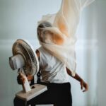 Kreatywny portret artystyczny osoby z welonem, wchodzącej w interakcję z wentylatorem elektrycznym w pomieszczeniu.