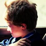 Młody chłopak siedzi w samochodzie i wygląda przez okno z zamyślonym wyrazem twarzy w świetle słońca.