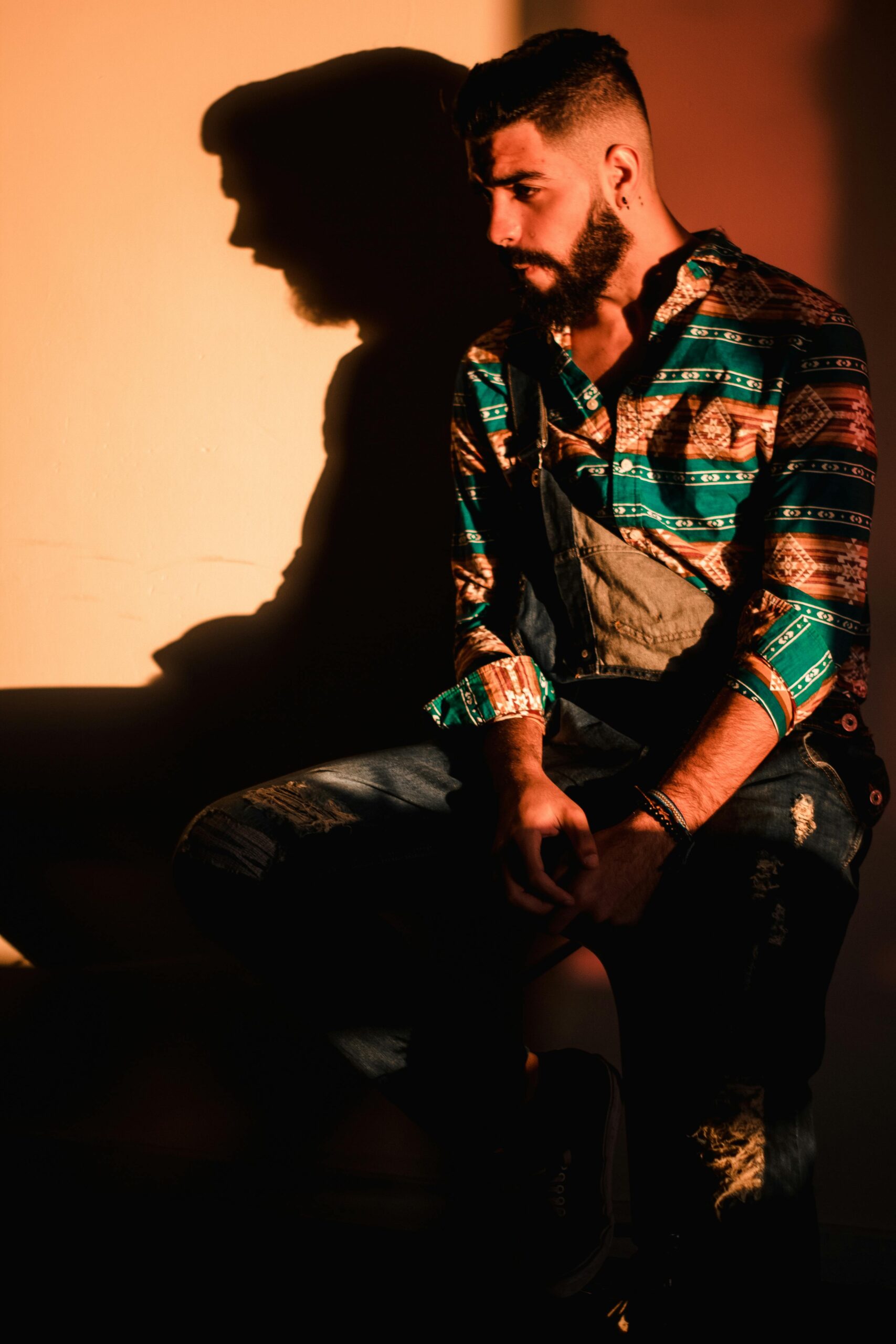 Modny mężczyzna w swobodnym stroju, siedzący w pomieszczeniu, z dramatyczną grą cieni.