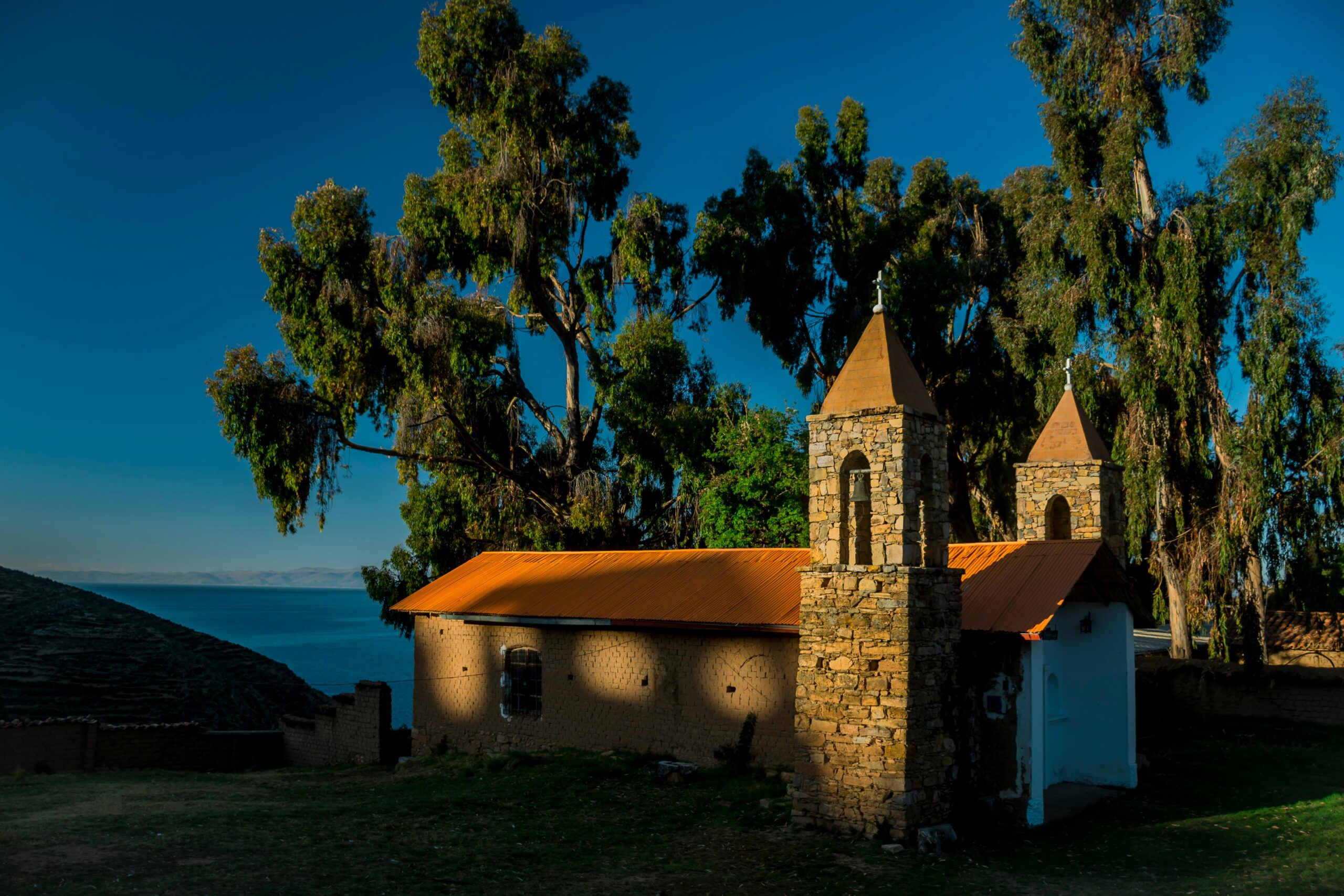 Urocza kamienna kaplica otoczona drzewami nad brzegiem jeziora Titicaca w ciepłym świetle słonecznym.