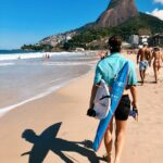 Surfer z deską spaceruje po plaży Ipanema w Rio de Janeiro, pod słonecznym niebem, na tle malowniczych gór.