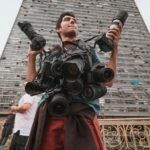 Mężczyzna trzyma wiele kamer, stojąc przed miejskim wieżowcem.