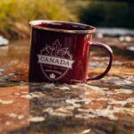 Czerwony metalowy kubek z wzorem Kanady umieszczony w jesiennej scenerii rzeki.