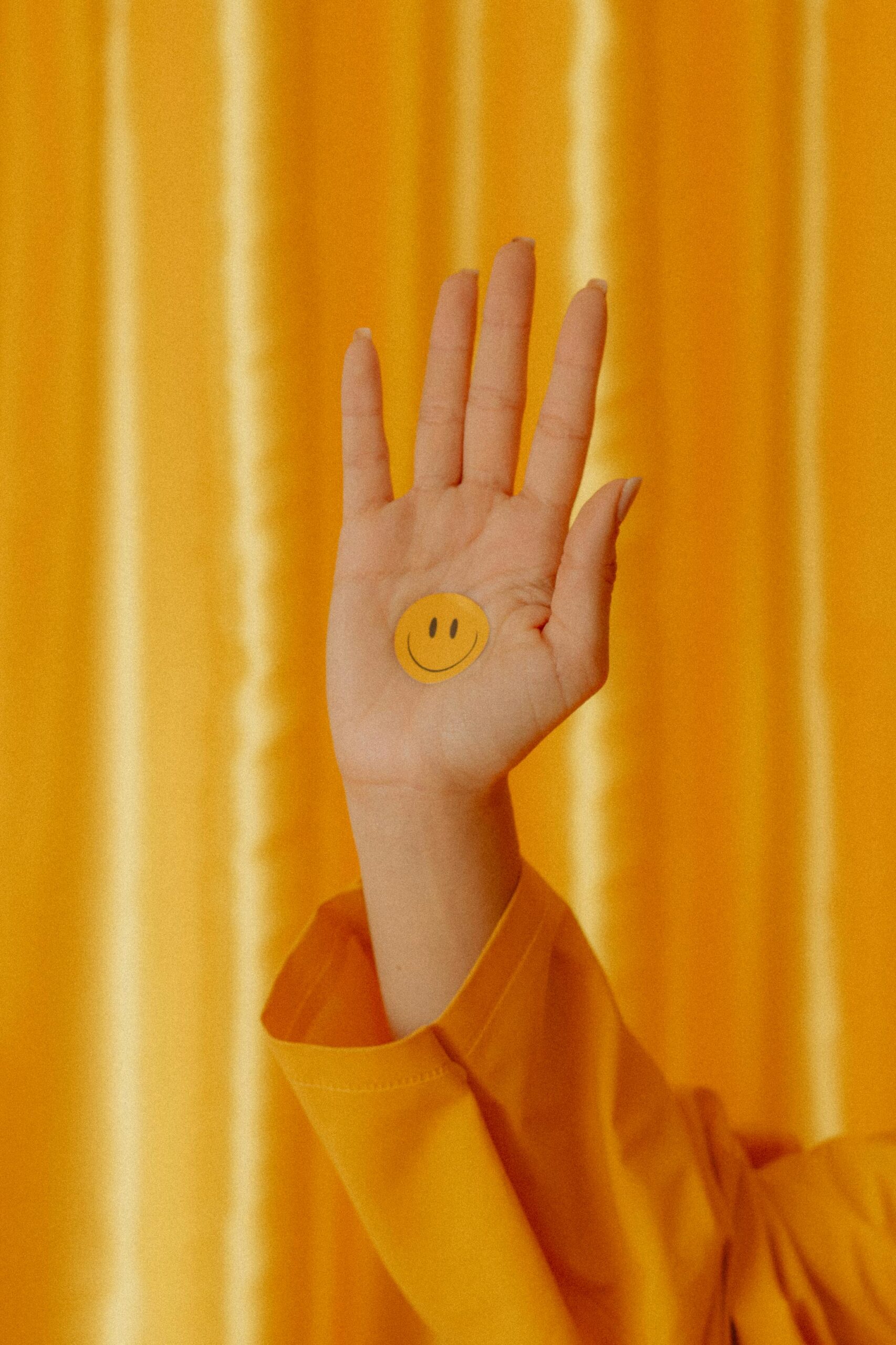 Dłoń z naklejką z uśmiechniętą buźką wyróżnia się na żywym żółtym tle, wyrażając pozytywność.