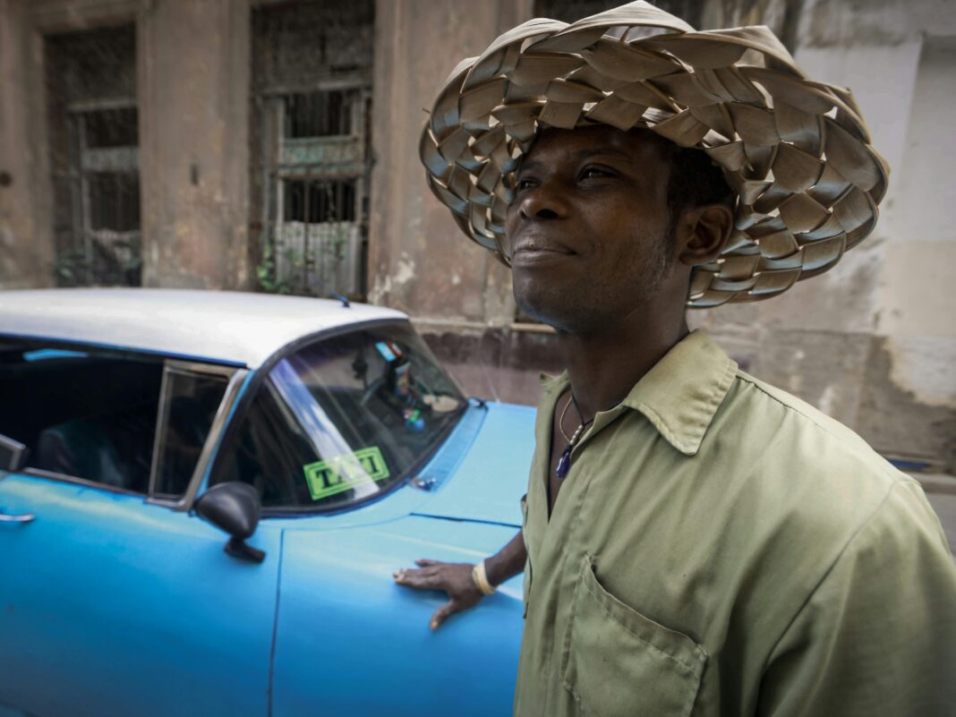 cuba Mężczyzna w tkanym kapeluszu stoi obok zabytkowej niebieskiej taksówki na ulicy miejskiej.