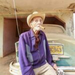 Starszy mężczyzna w kapeluszu i kurtce pali cygaro, siedząc przy zabytkowym samochodzie na Kubie.