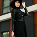 Modna kobieta w czarnym stroju i kapeluszu pewnie pozuje na świeżym powietrzu w miejskim otoczeniu, prezentując nowoczesny styl.