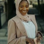 Portret uśmiechniętej kobiety w różowym hidżabie i płaszczu, trzymającej torbę na zewnątrz.