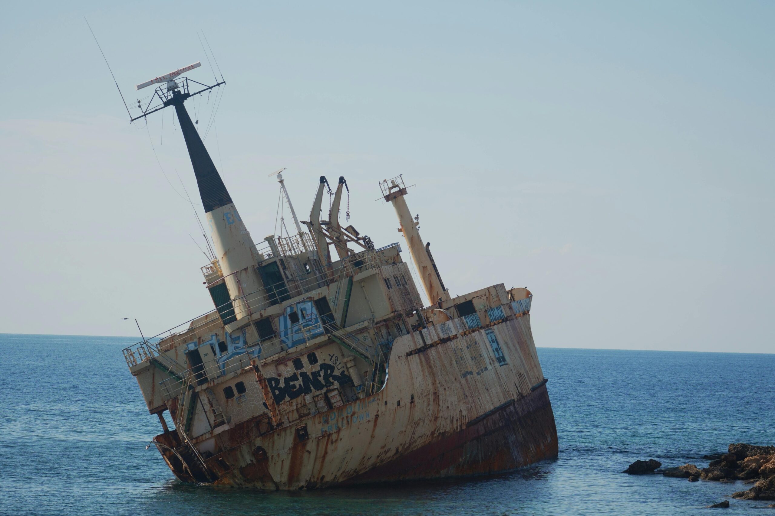 Opuszczony zardzewiały wrak statku pochylony w stronę morza w spokojny, słoneczny dzień w pobliżu skalistych brzegów.