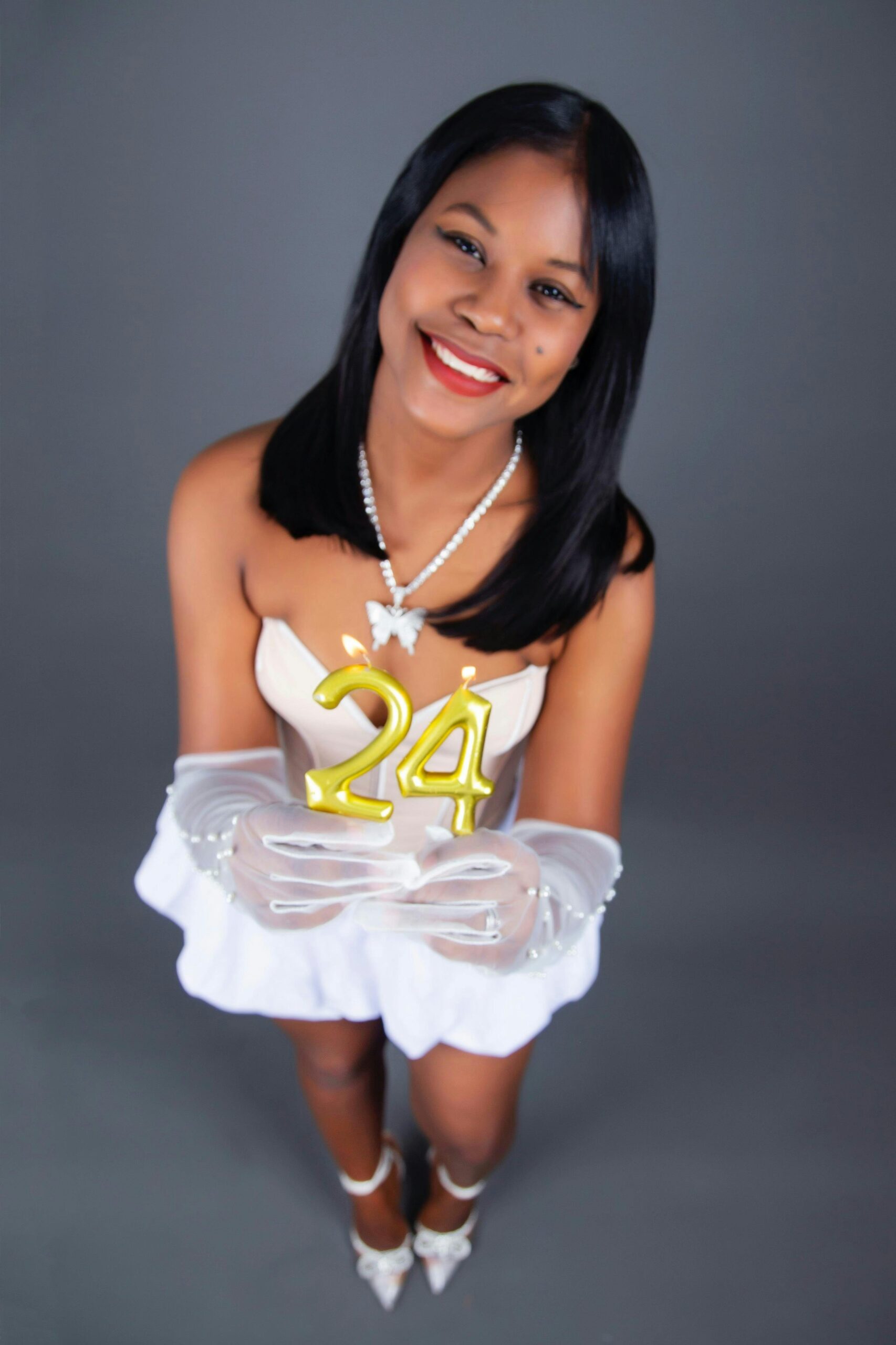 Kobieta w białej sukni trzymająca topper na tort „24”, uśmiechając się radośnie w eleganckich rękawiczkach.