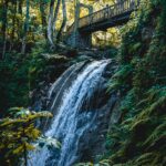 Piękny wodospad spływający kaskadą pod drewnianym mostem otoczonym bujną roślinnością w leśnej scenerii.