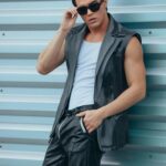 Model płci męskiej w skórze i okularach przeciwsłonecznych pewnie pozuje na zewnątrz na metalowym tle.