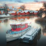 Finlandia latem – jeziora, sauny i niezapomniane krajobrazy Pokryte śniegiem łodzie odbijające się w historycznym porcie Porvoo podczas zimowego zachodu słońca.