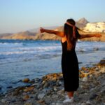 Antyczne Skarby Aten: Przegląd Najważniejszych Zabytków Stolicy Grecji i Opowieść o Ich Historii Kobieta ciesząca się wolnością o zachodzie słońca na skalistej plaży w Grecji.