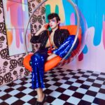 Stylowa osoba pozuje w pokoju w stylu retro, z odważnymi kolorami i wzorami, ubrana w cekinowy strój.