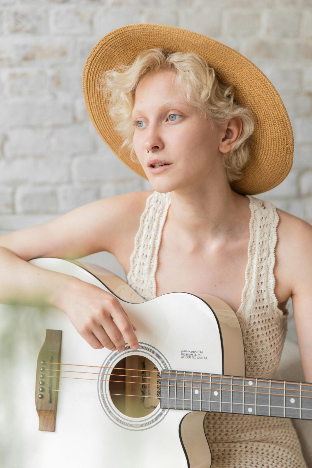 Młoda kobieta w słomkowym kapeluszu i letniej sukience gra na gitarze w pomieszczeniu z pogodnym wyrazem twarzy.