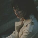 Spokojny portret kobiety w trenczu, siedzącej na zewnątrz z rozwianymi włosami.