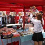 Kobieta organizuje wydruki artystyczne na tętniącym życiem straganie z kolorowymi ubraniami i dodatkami.