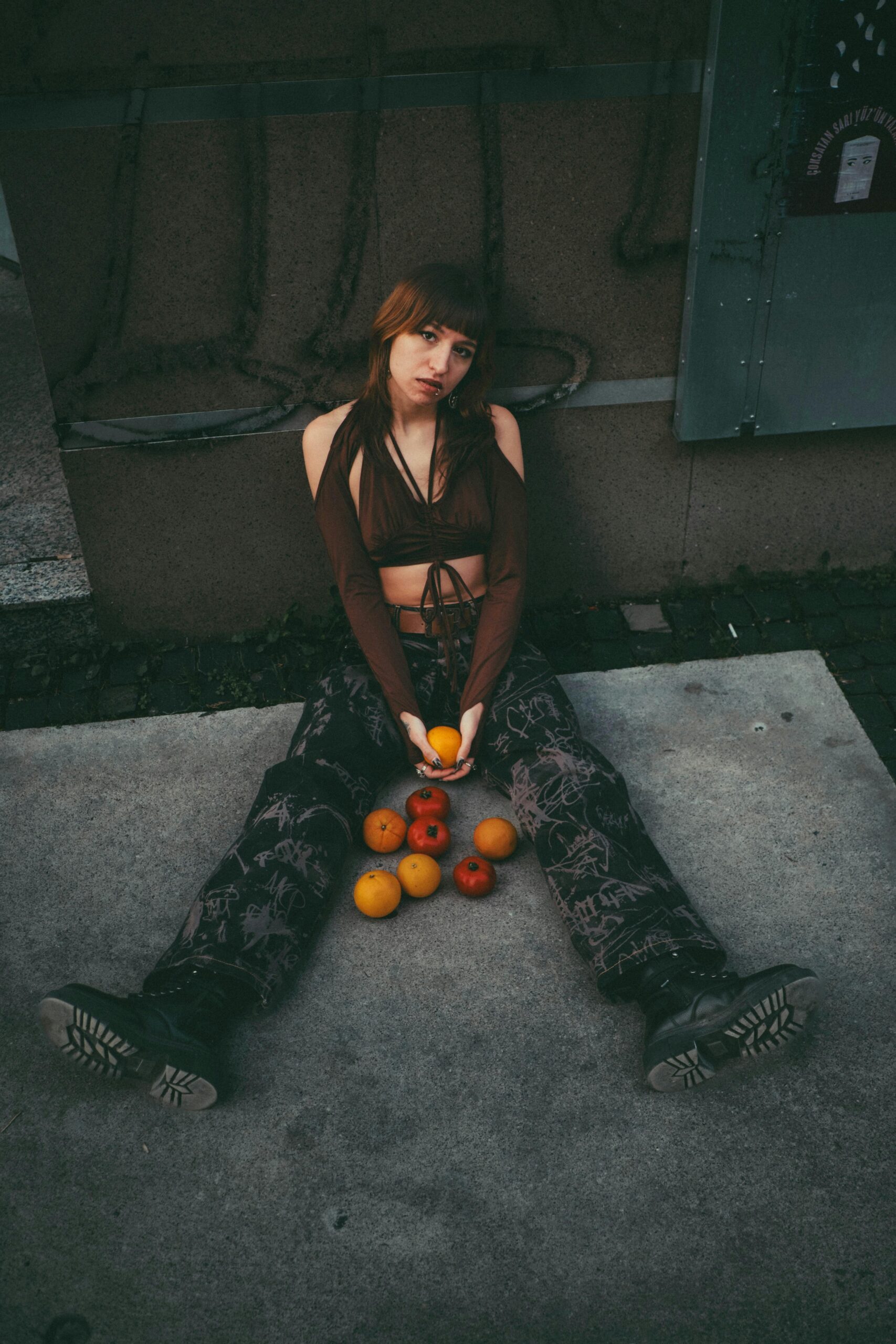 Artystyczny portret kobiety siedzącej na ziemi z porozrzucanymi pomarańczami i jabłkami, emanującej nastrojową atmosferą.