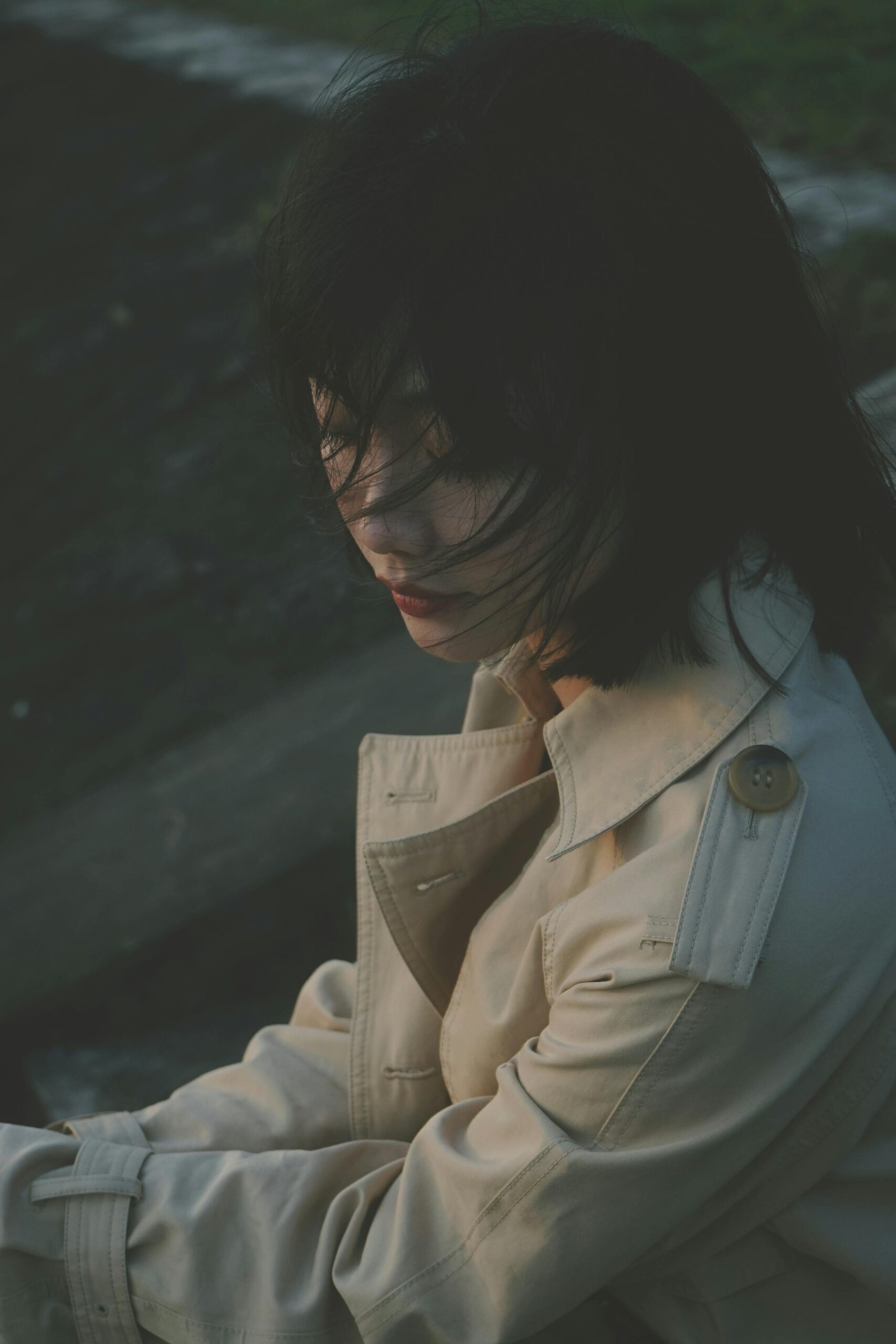 Spokojny portret kobiety w trenczu, siedzącej na zewnątrz z rozwianymi włosami.