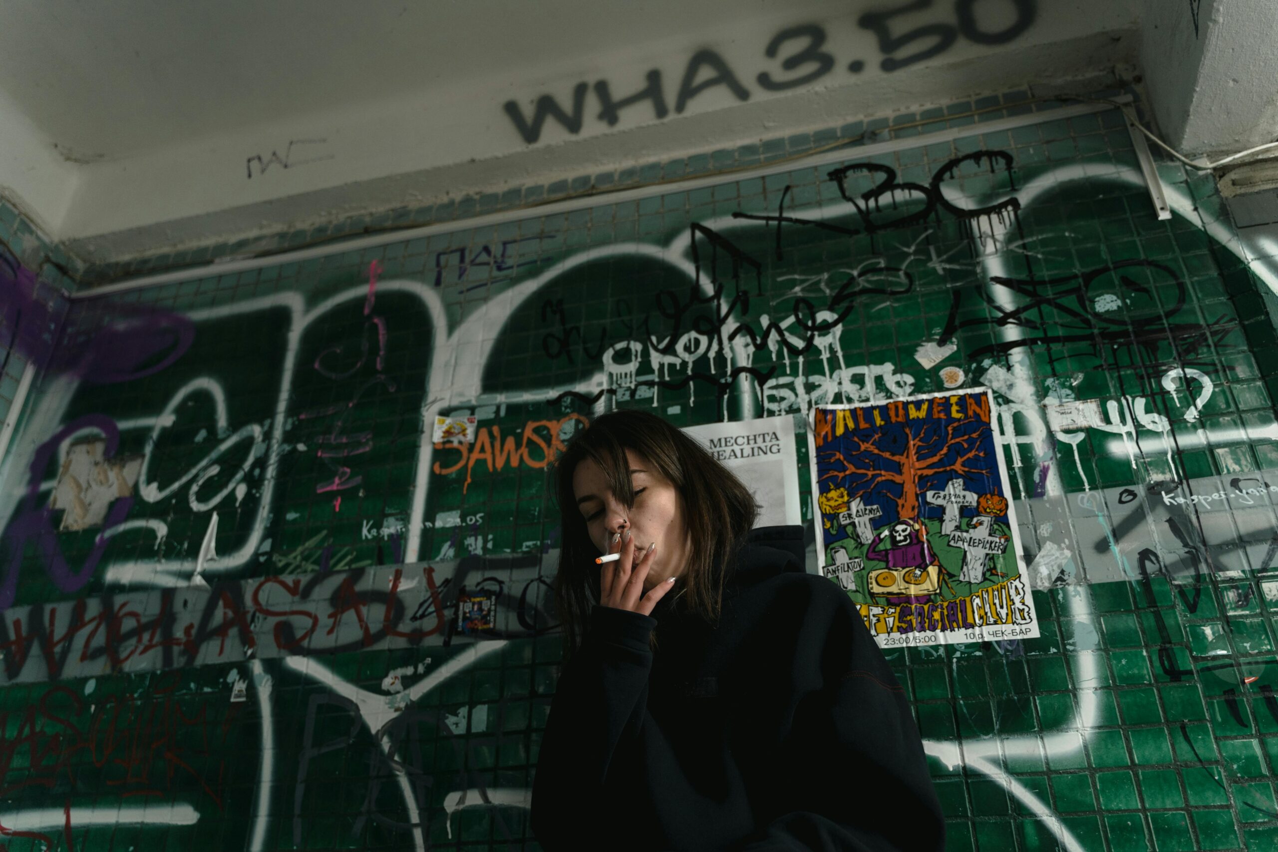 Kobieta w czarnej bluzie z kapturem pali przy ścianie pokrytej graffiti w miejskim otoczeniu.