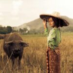 Kobieta na wiejskich terenach Indonezji stoi na polu ryżowym z bawołem, ubrana w tradycyjny strój.